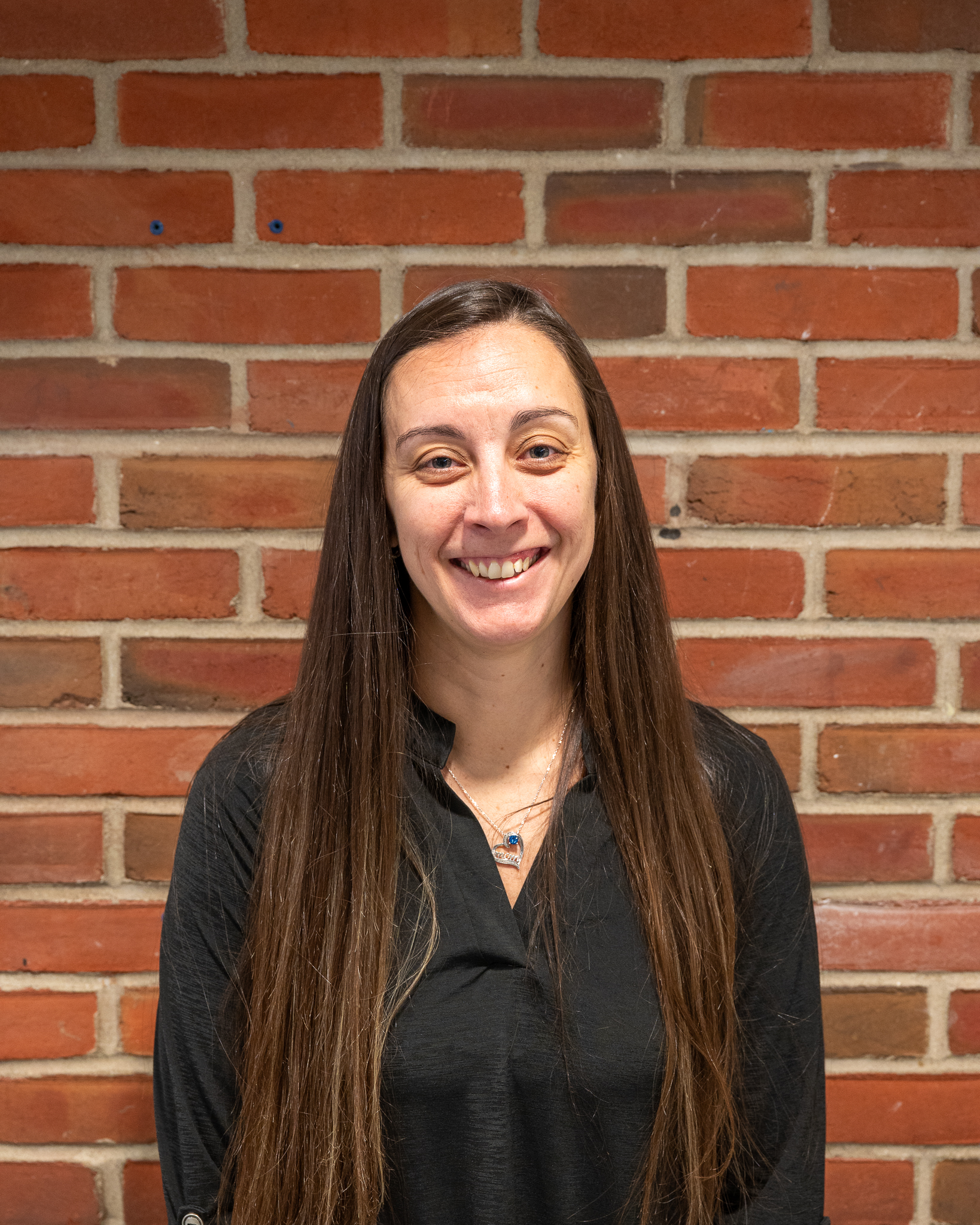 A woman with long brown hair stands smiling against a red brick wall.